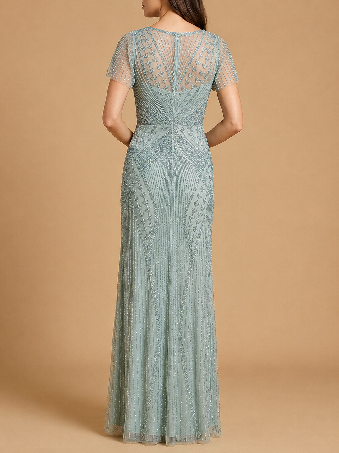 A woman modeling a floor-length, short-sleeve evening gown from the back, featuring intricate beadwork and lace detailing in a dusty sage green color, set against a solid tan studio background.